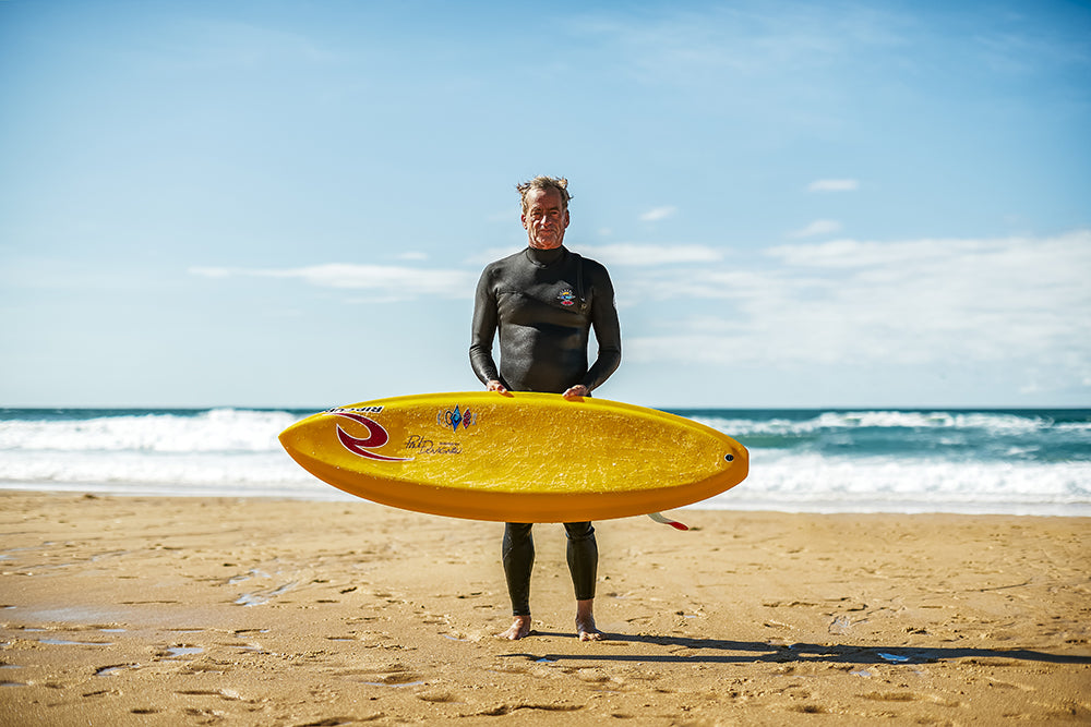 Person in a wetsuit holding a yellow surfboard on a beach with ocean waves in the background