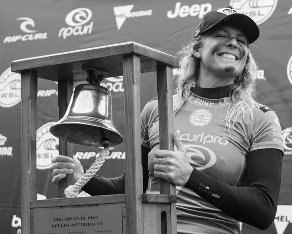 Person in a wetsuit holding a bell with event branding in the background