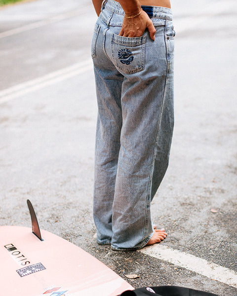 Person wearing light blue jeans standing on a road with a pink surfboard nearby.