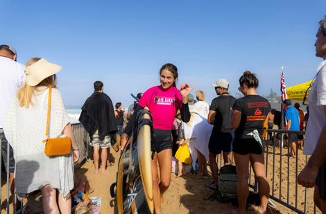Woman in pink shirt and black shorts walking on a sandy beach with people around her.