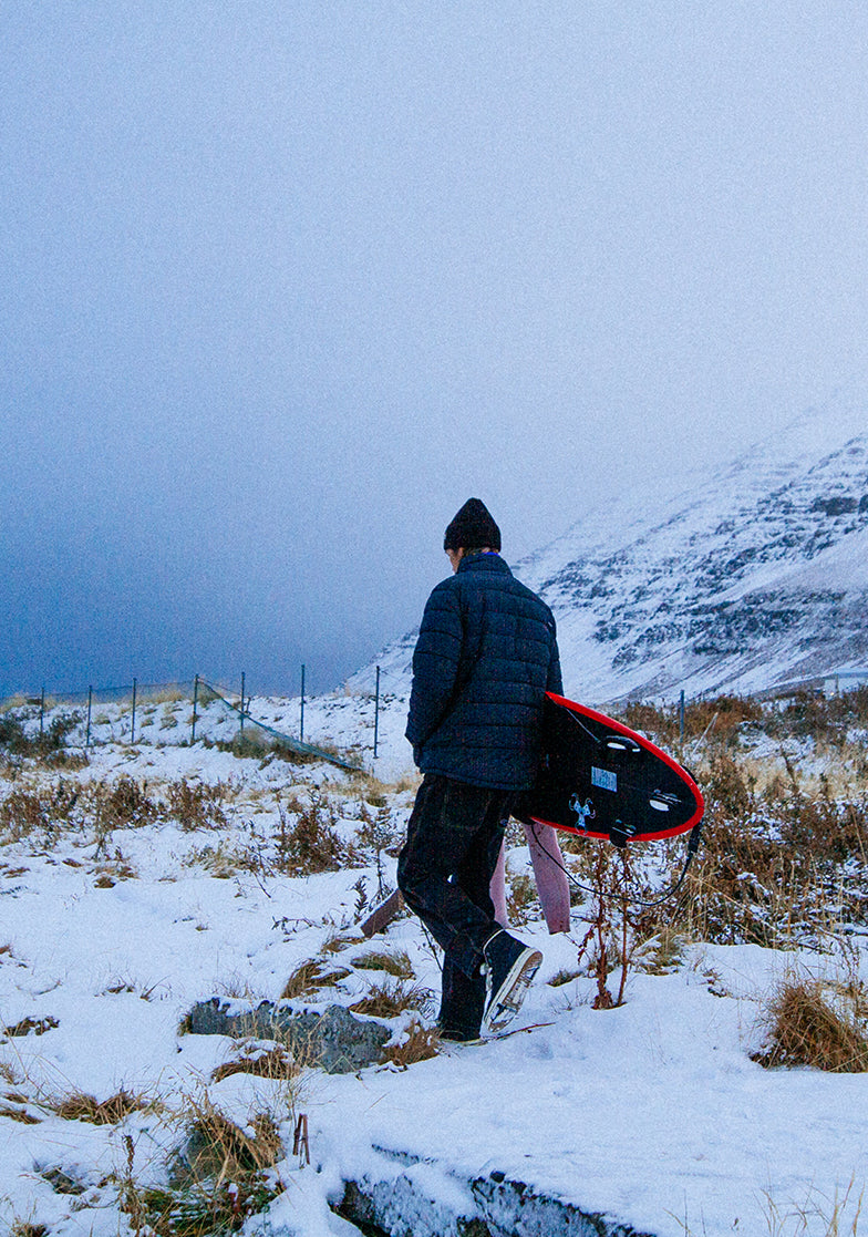 Person walking with a surfboard on a snowy mountain path