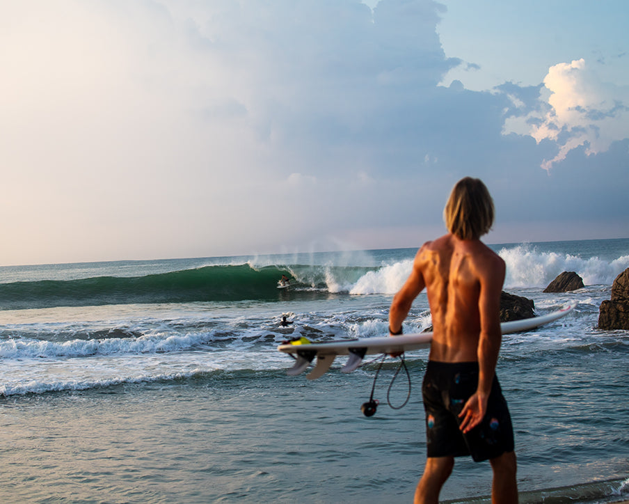 Man with a surfboard standing on a beach looking at waves