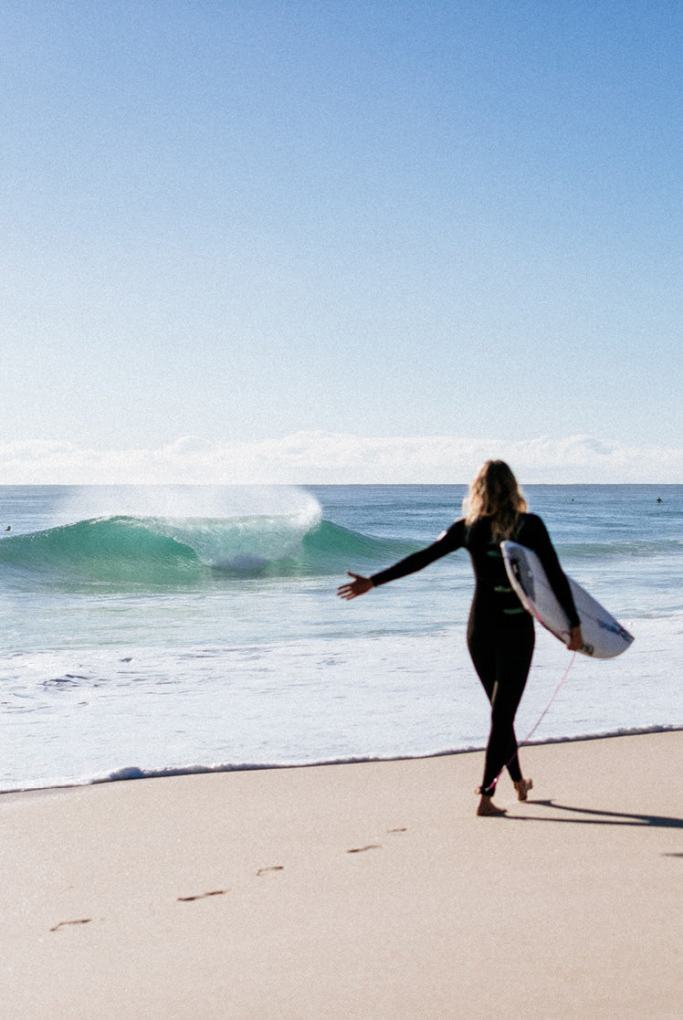 Person in a wetsuit holding a surfboard on a beach with waves in the background