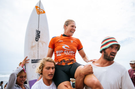 Person in orange Rip Curl shirt being carried by others with a surfboard in the background